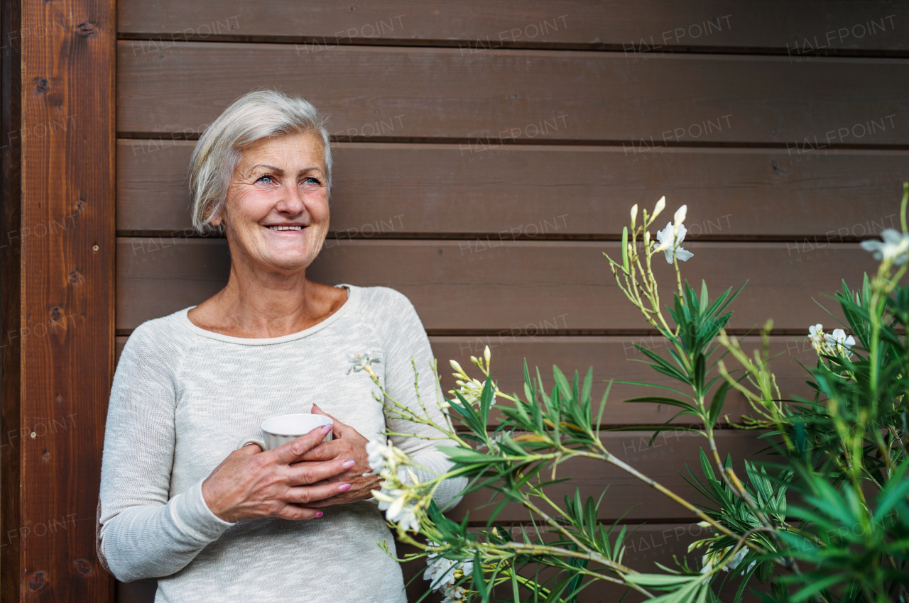 A happy senior woman with coffee standing outdoors on a terrace in summer.