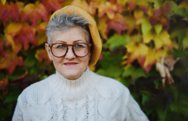 Portrait of senior woman standing outdoors against colorful natural autumn background, looking at camera.