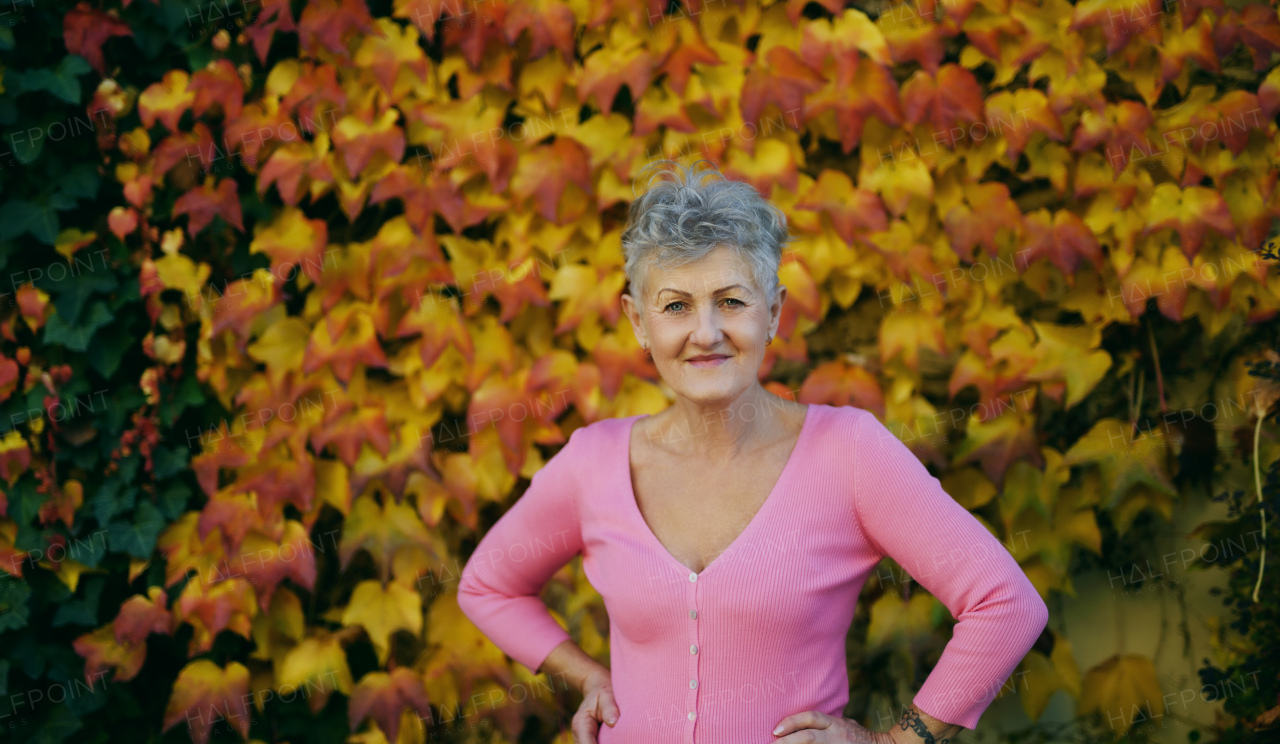 Portrait of senior woman standing outdoors against colorful natural autumn background, looking at camera.