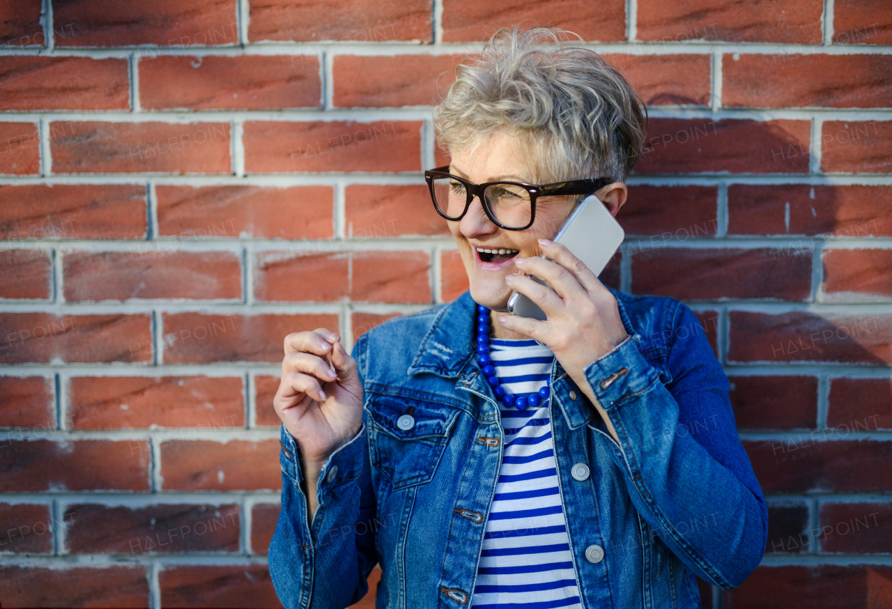 Portrait of senior woman standing outdoors against brick wall background, using smartphone.