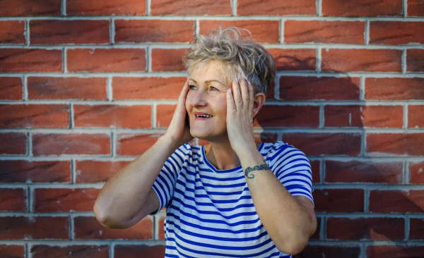 Portrait of senior woman standing outdoors against brick wall background, relaxing.