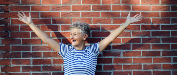 Portrait of senior woman standing outdoors against brick wall background, stretching arms.