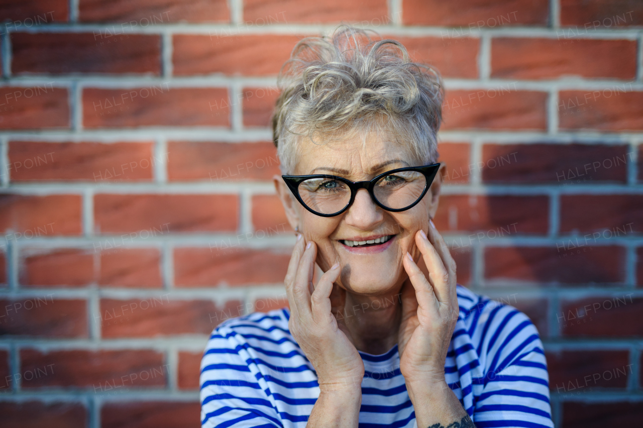 Portrait of senior woman standing outdoors against brick wall background, looking at camera.