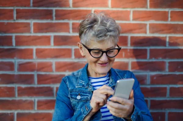 Portrait of senior woman standing outdoors against brick wall background, using smartphone.