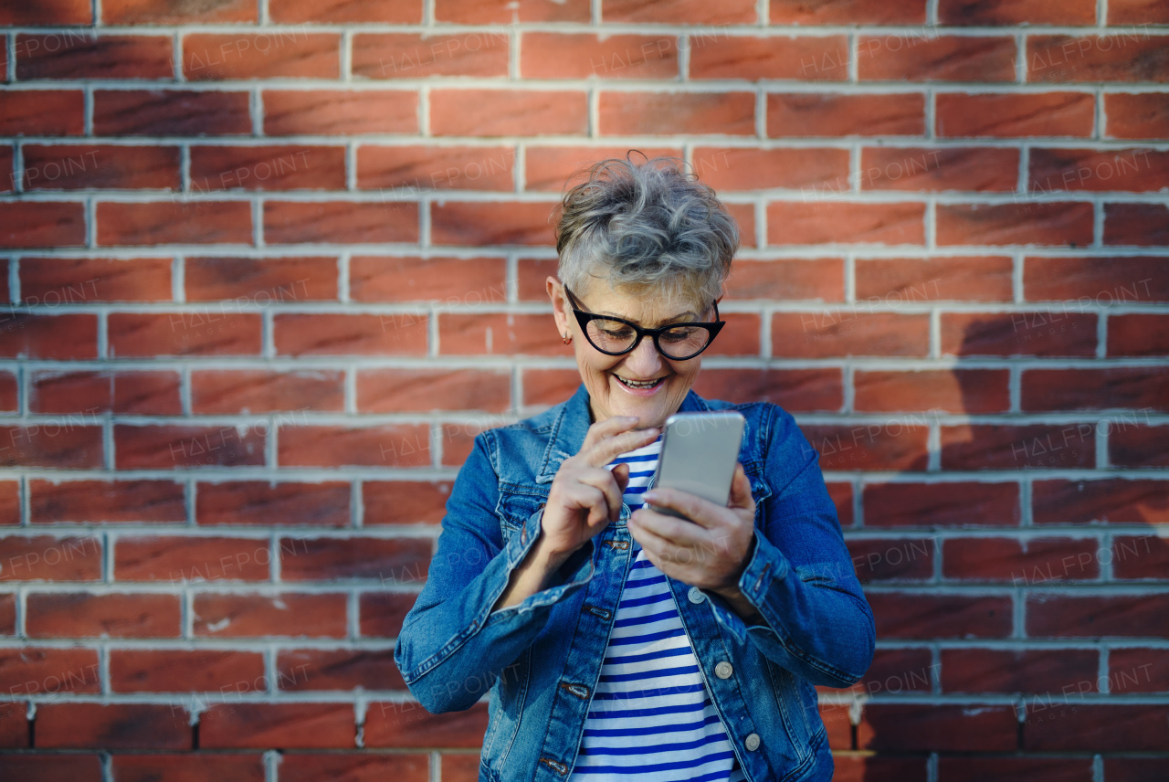 Portrait of senior woman standing outdoors against brick wall background, using smartphone.