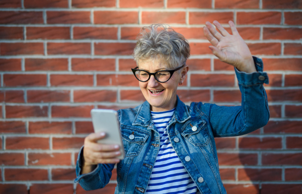 Portrait of senior woman standing outdoors against brick wall background, using smartphone for video call.