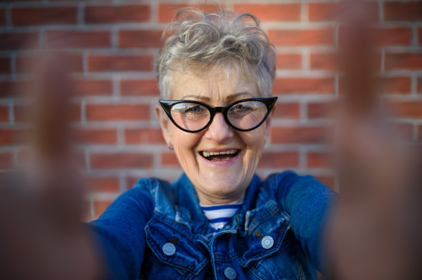 Portrait of senior woman standing outdoors against brick wall background, taking selfie.