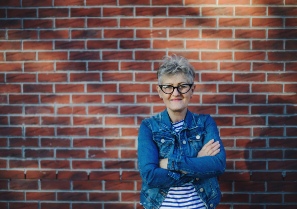 A senior woman standing outdoors against brick wall background, looking at camera.