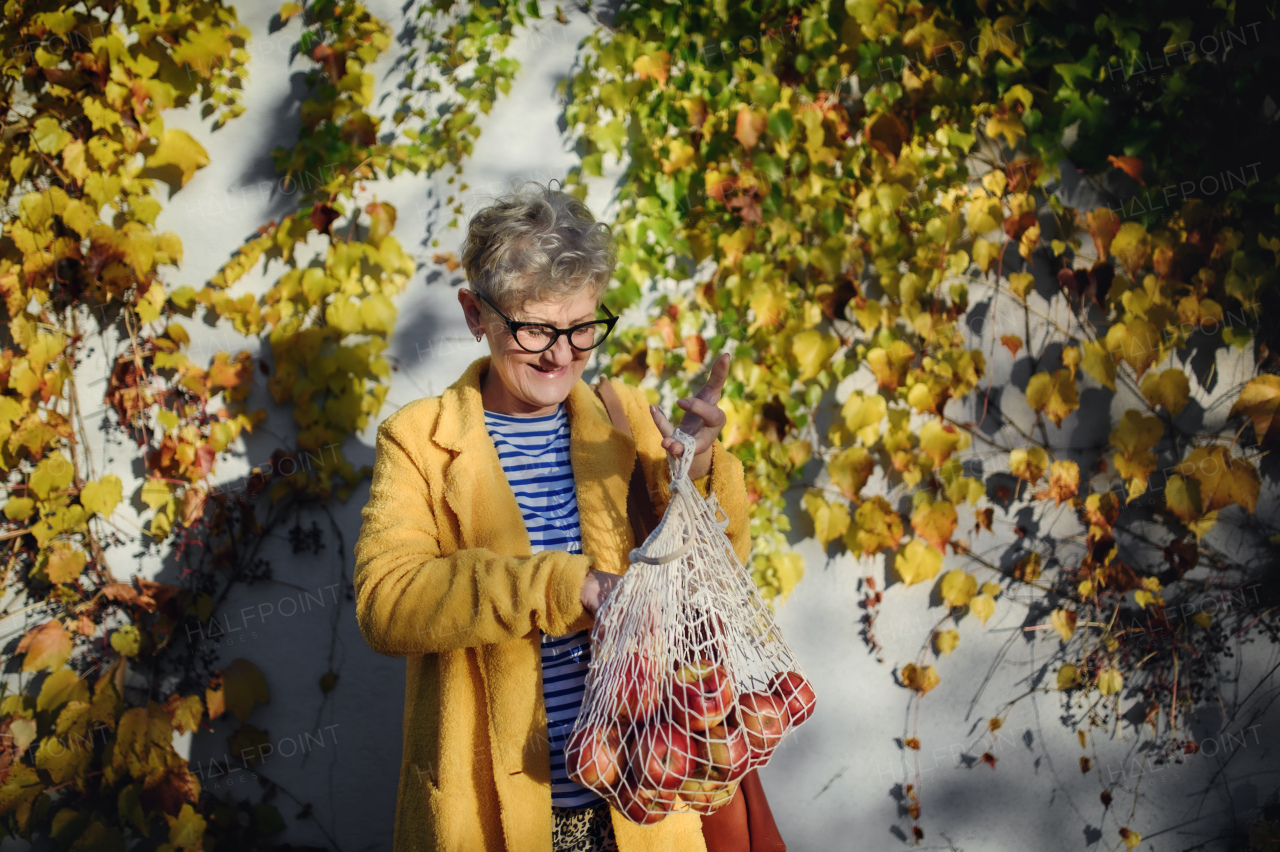 Senior woman standing outdoors against colorful natural autumn background, holding shopping in reusable bag.