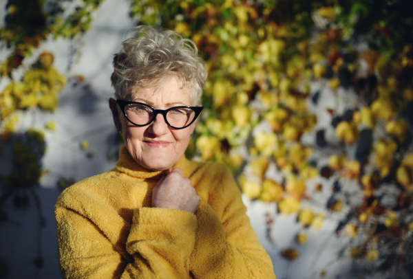 Portrait of senior woman standing outdoors against wall in town, looking at camera.