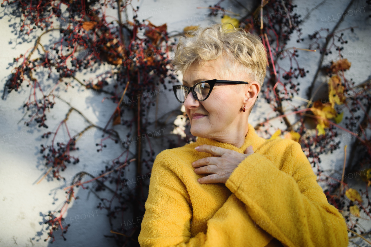 Portrait of senior woman standing outdoors against wall in town.