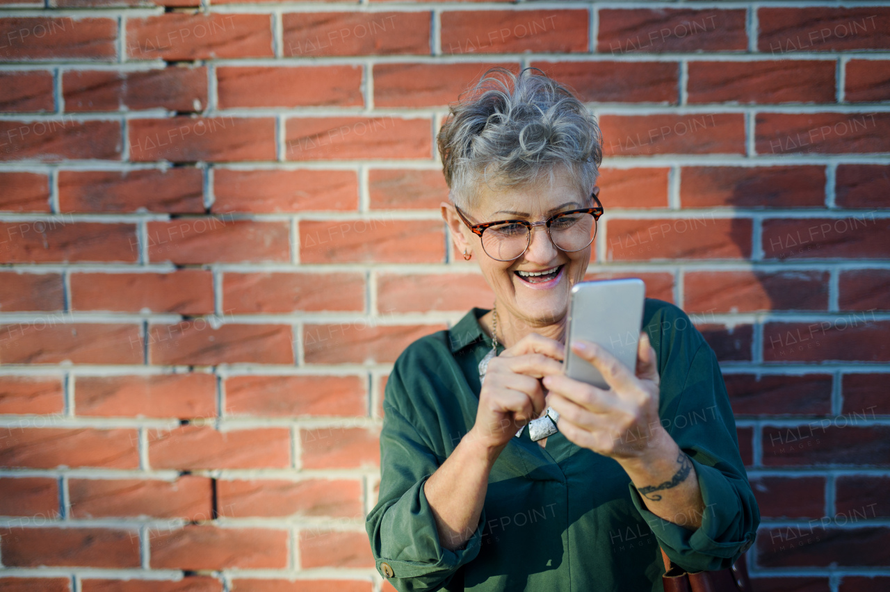 Portrait of senior woman standing outdoors against brick wall background, using smartphone.