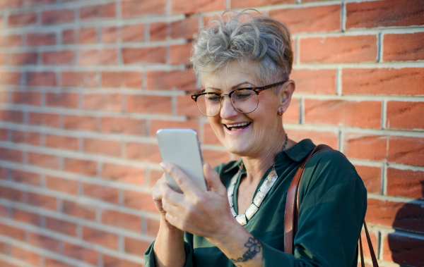 Portrait of senior woman standing outdoors against brick wall background, using smartphone.