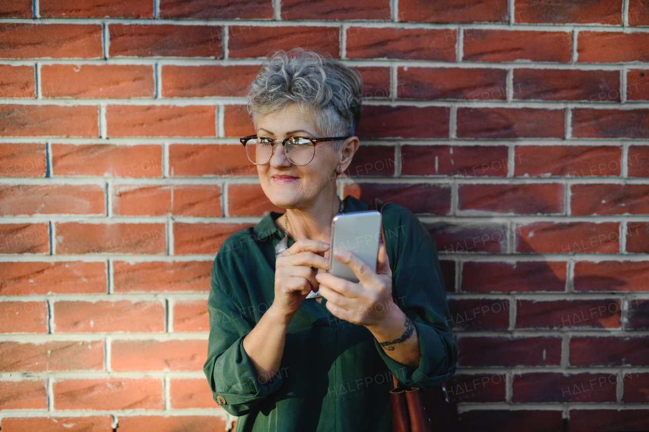 Portrait of senior woman standing outdoors against brick wall background, using smartphone.