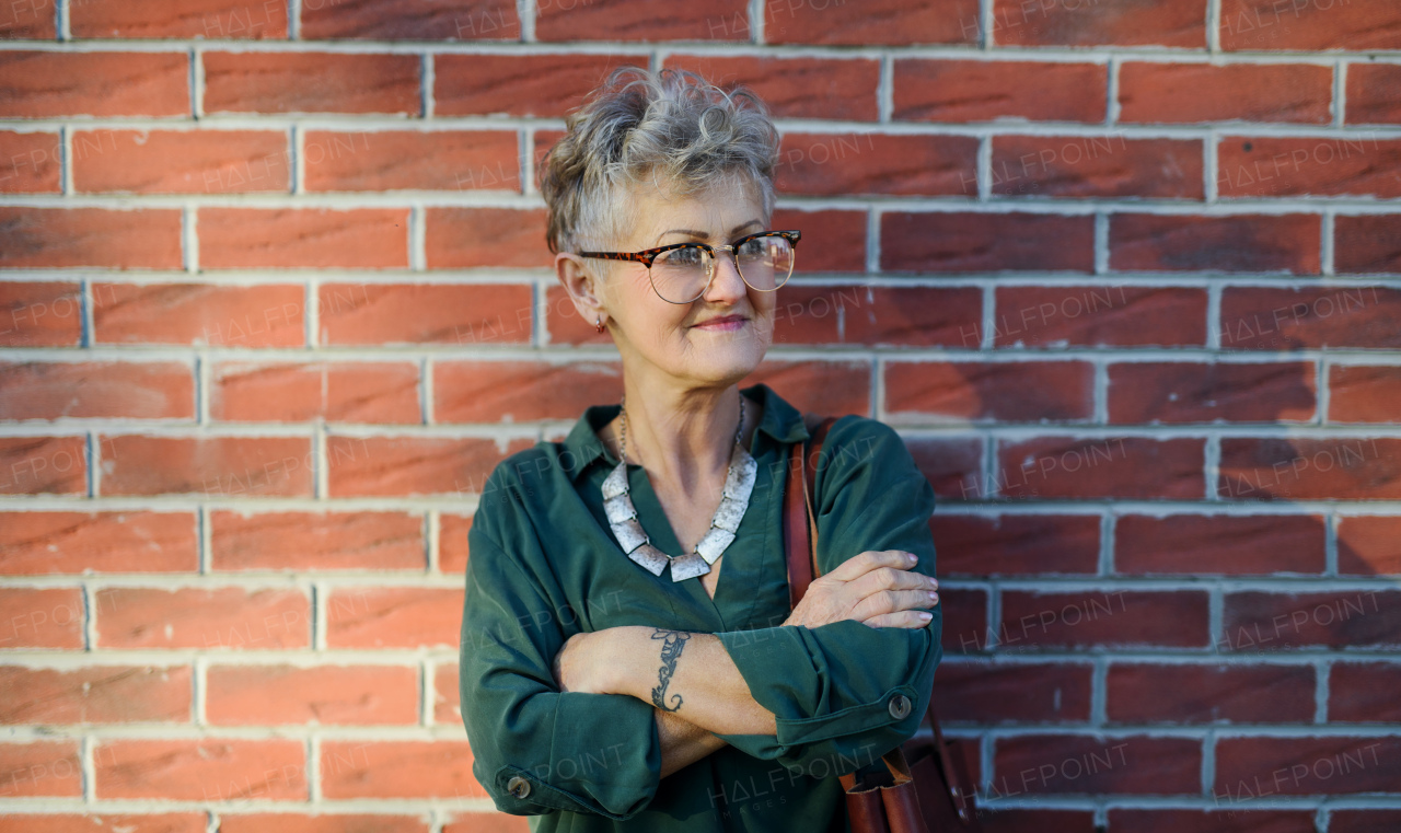 Portrait of senior woman standing outdoors against brick wall in town, arms crossed.