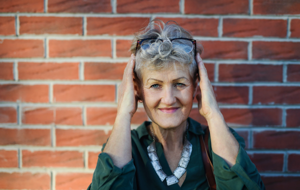 Portrait of senior woman standing outdoors against brick wall in town, looking at camera.