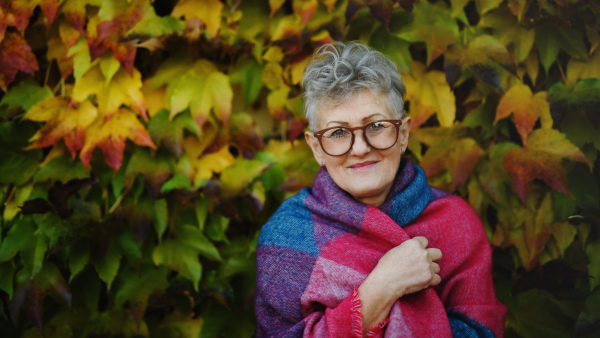 Portrait of senior woman standing outdoors against colorful natural autumn background, looking at camera.