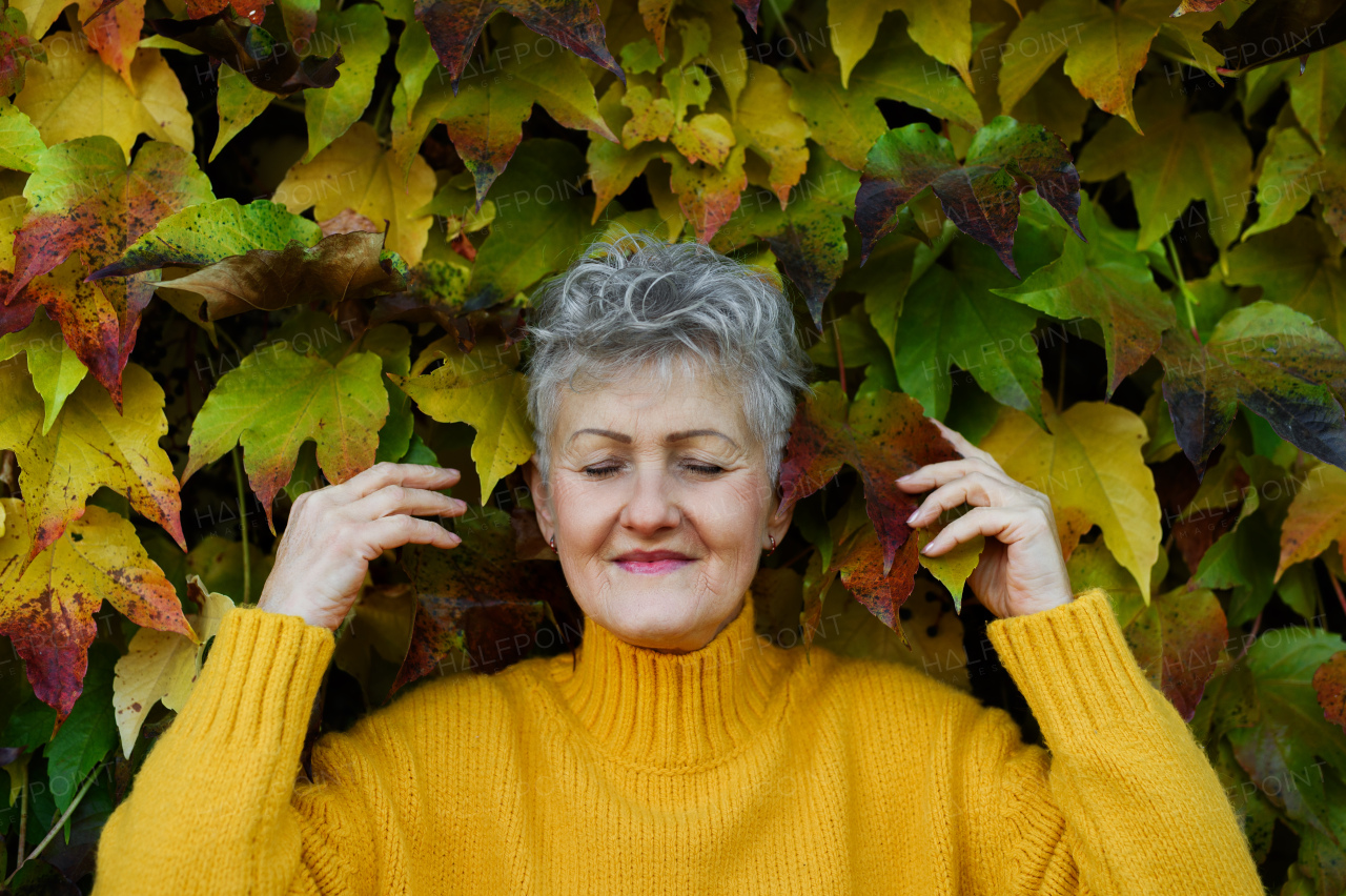 Portrait of senior woman standing outdoors against colorful natural autumn background, eyes closed.