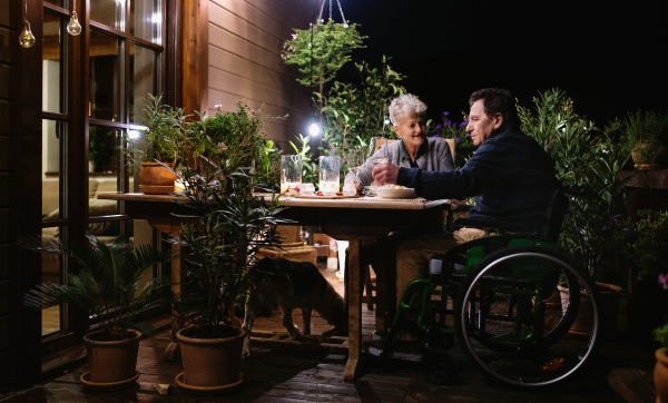 Happy senior couple in wheelchair having dinner in the evening on terrace, drinking wine.