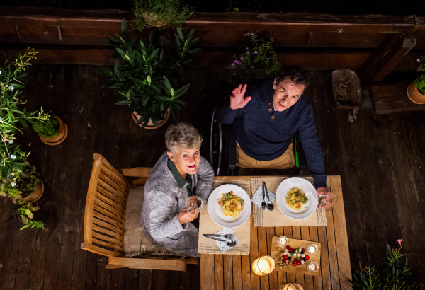 Top view of senior couple in wheelchair having dinner in the evening on terrace, looking at camera.