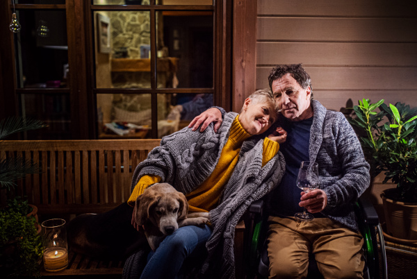 Portrait of senior couple in wheelchair with dog in the evening on terrace, drinking wine.