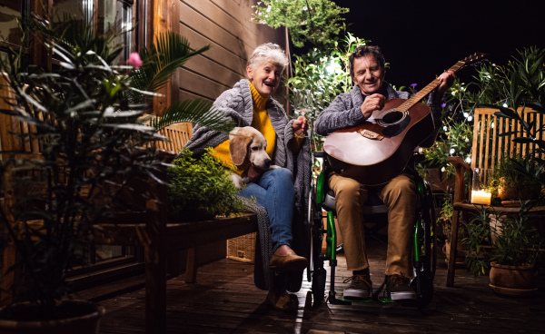 Senior couple in wheelchair with dog and guitar in the evening on terrace, resting.