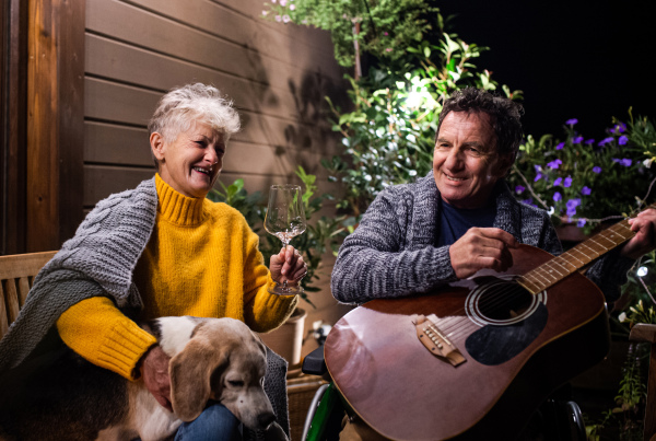 Senior couple in wheelchair with dog and guitar in the evening on terrace, resting.