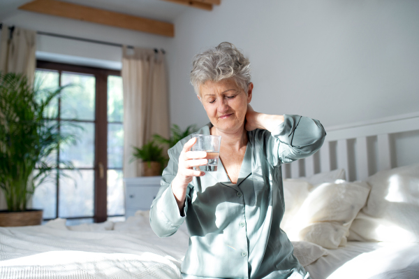 A senior woman with neck pain getting up from bed in the morning, health problems concept.