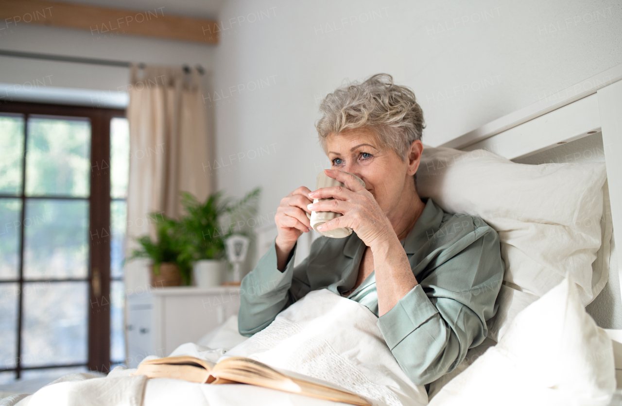 Happy senior woman reading a book and drinking coffee in bed at home.