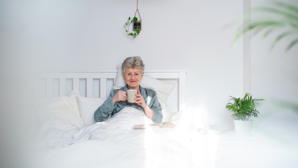 Portrait of happy senior woman with coffee reading book in bed at home, looking at camera.