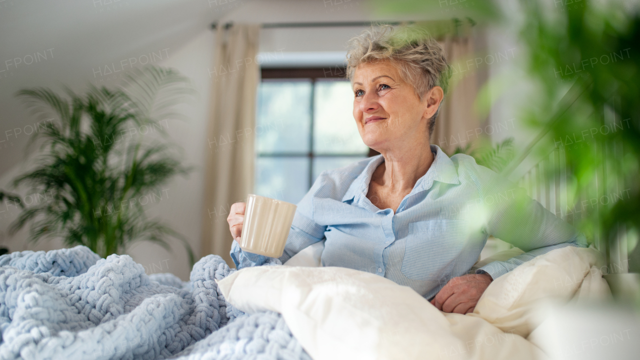 A happy senior woman with a hot drink in bed at home, relaxing.