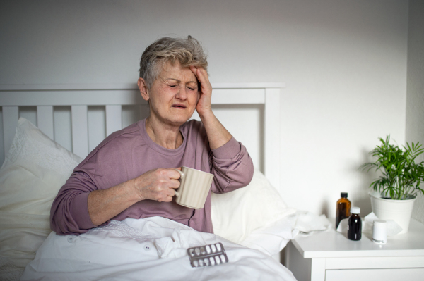 A sick senior woman in bed at home, headache, illness and taking medication concept.