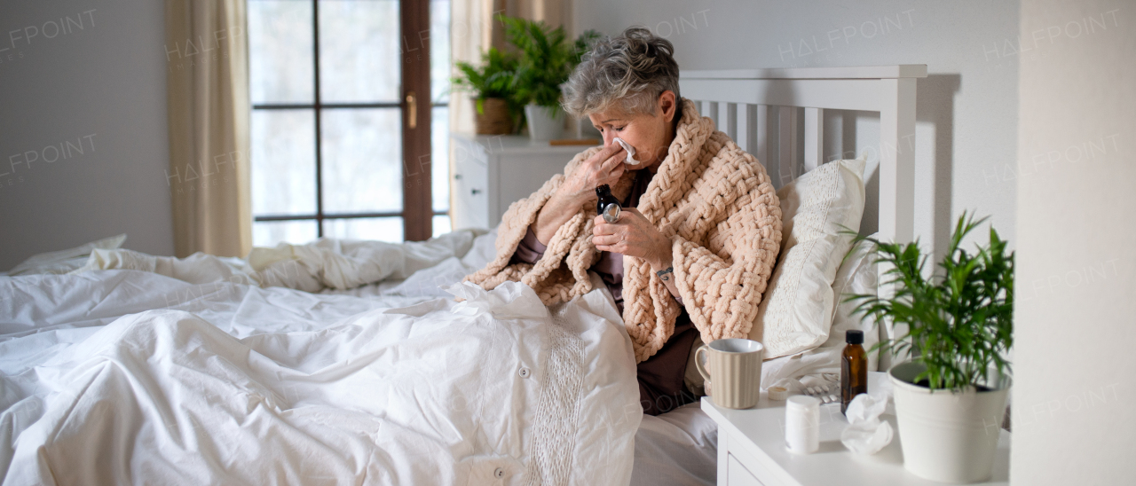 A sick senior woman in bed at home, sneezing and taking medication.