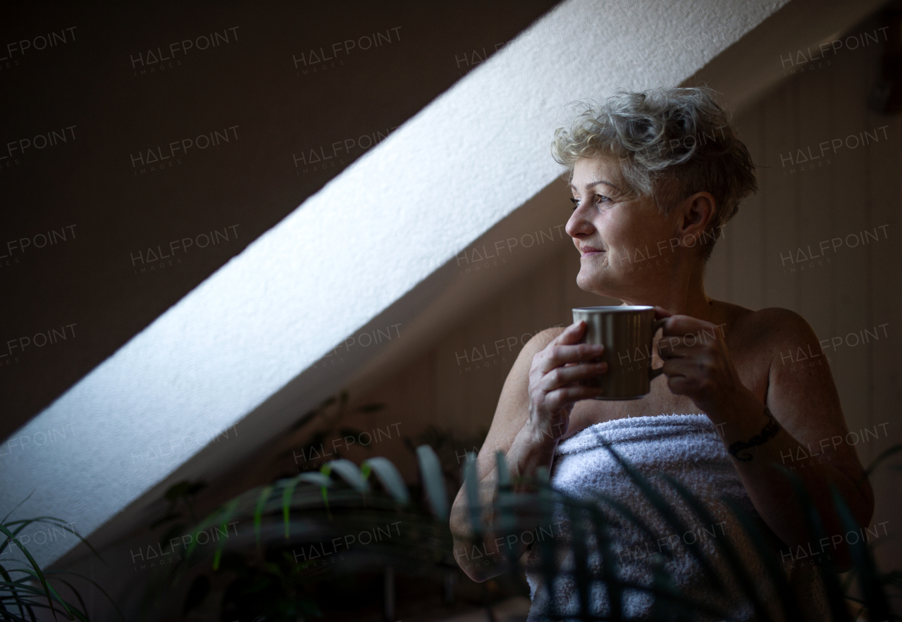 Happy senior woman wrapped in a towel in bathroom at home, sitting and resting with tea.