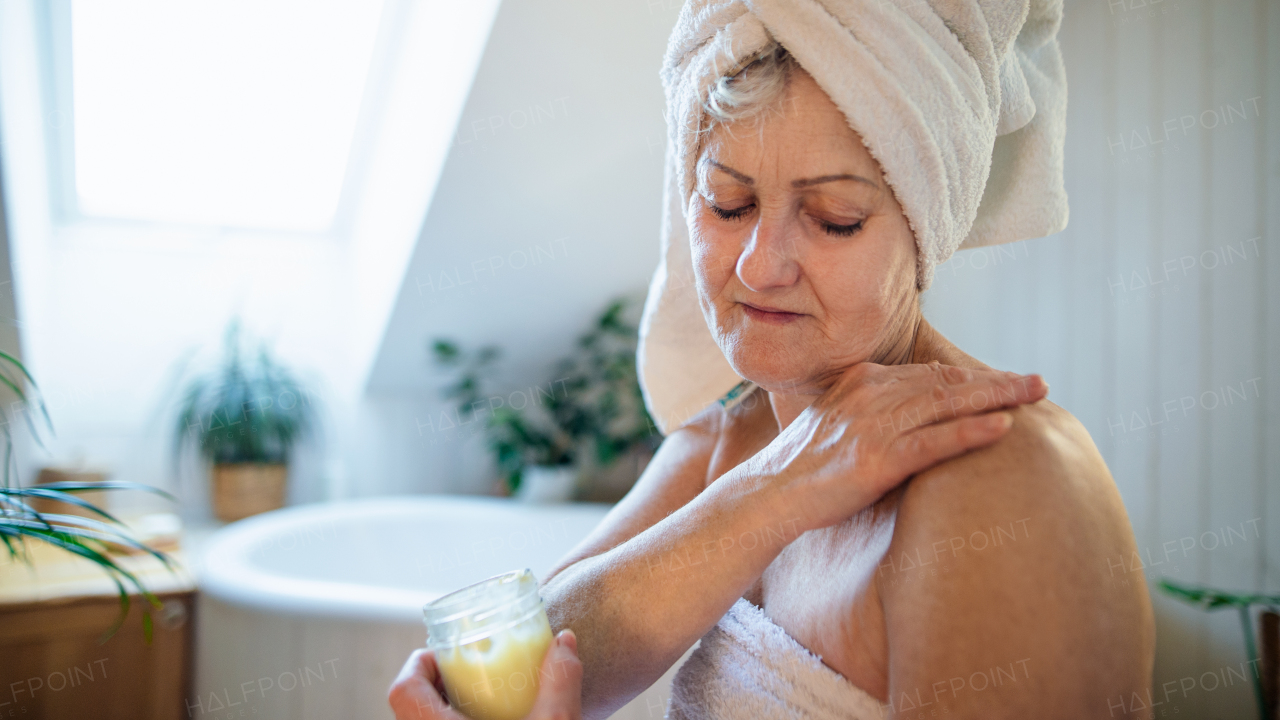 A happy senior woman wrapped in a towel in bathroom at home, applying homemade body moisturiser.