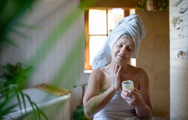 A happy senior woman wrapped in a towel in bathroom at home, applying homemade body moisturiser.