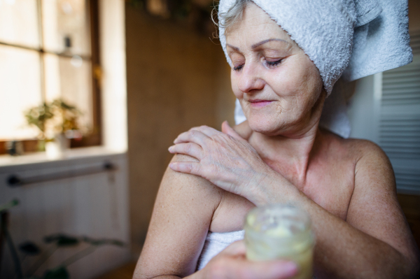 A happy senior woman wrapped in a towel in bathroom at home, applying homemade body moisturiser.