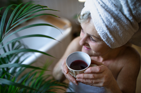 Happy senior woman wrapped in a towel in bathroom at home, sitting and resting with tea.