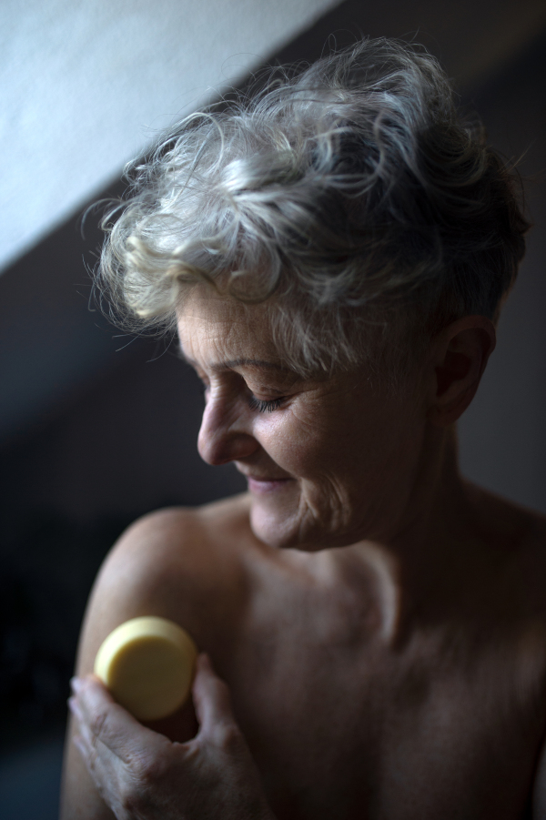 Portrait of happy topless senior woman in bathroom at home, using massage cube.