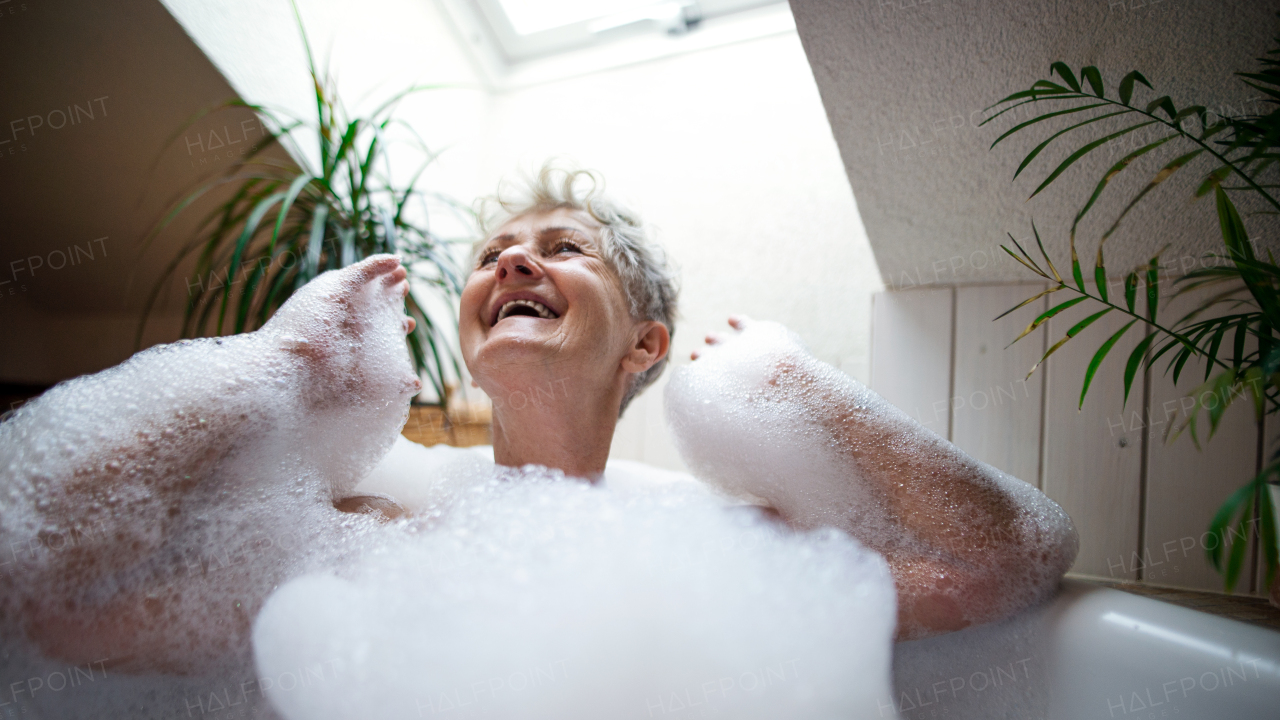 Portrait of cheerful senior woman washing in bubble bath tub at home, laughing.