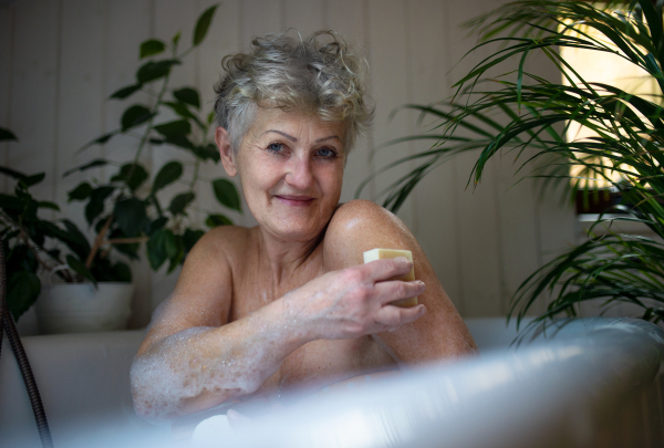 Portrait of contented senior woman washing in bubble bath tub at home, looking at camera.