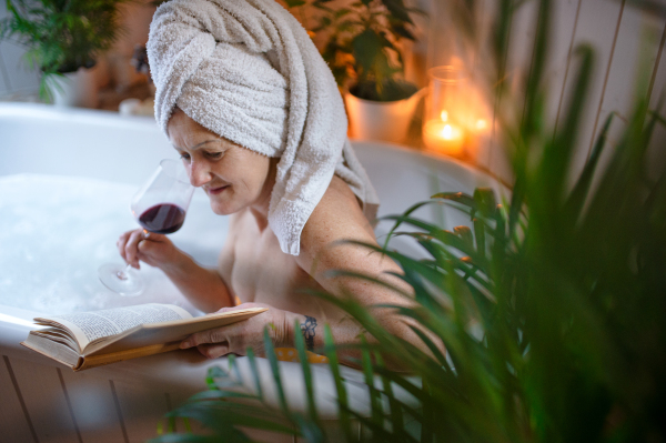 A happy senior woman reading book and drinking wine in bath tub at home.