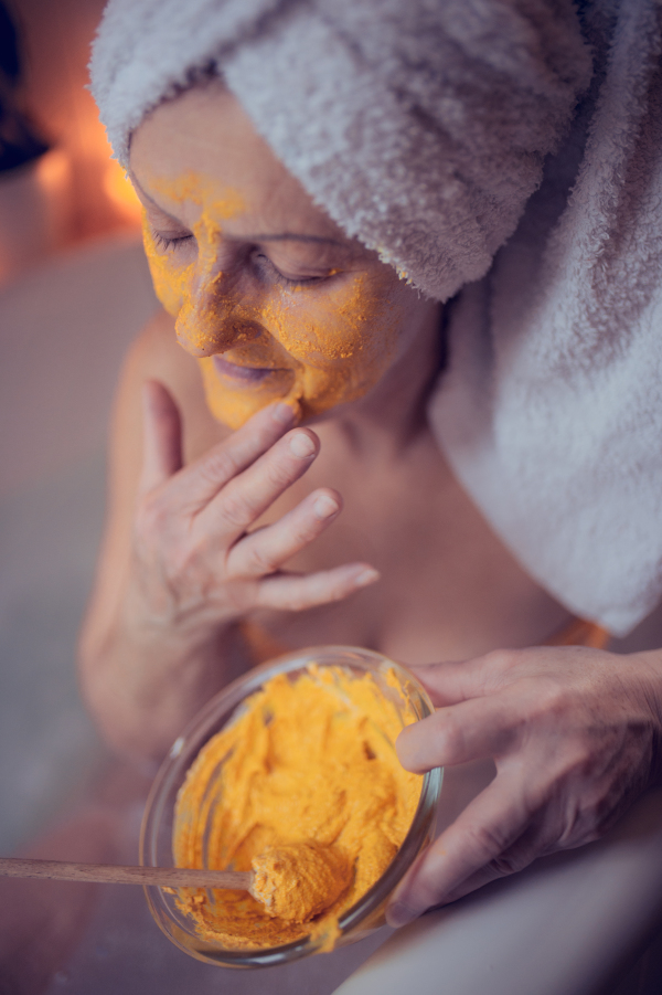 Happy senior woman applying beauty face mask in bath tub at home, relaxing with eyes closed.