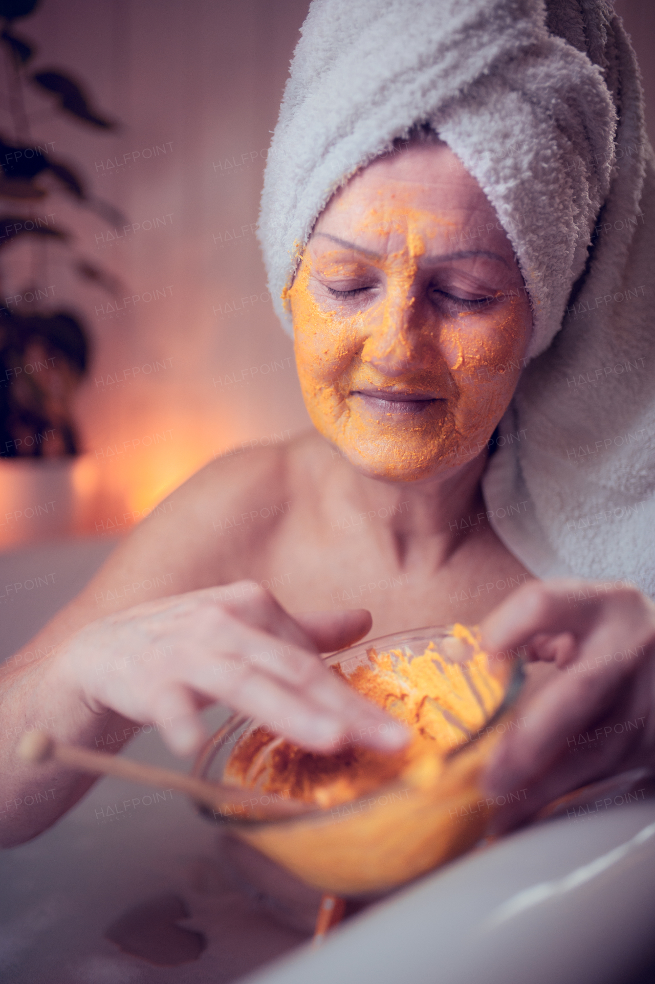 Happy senior woman applying beauty face mask in bath tub at home, relaxing with eyes closed.