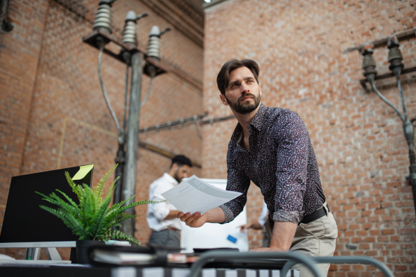 A thoughtful young businessman standing at desk in office, working.