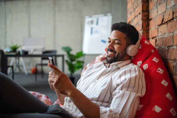 A cheerful young businessman with headphones and smartphone in office, taking a break and relaxing.