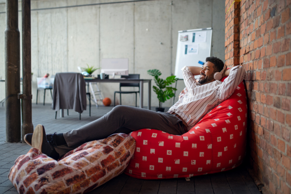 A cheerful young businessman with headphones lying on bean bag in office, taking a break and relaxing.