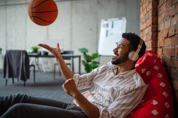 A cheerful young businessman with headphones and ball taking a break in office, resting.