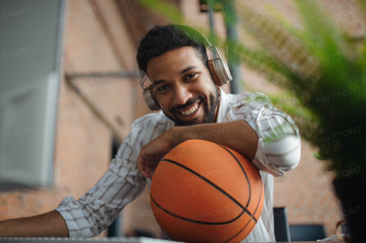 A cheerful young businessman with headphones and ball taking a break in office, looking at camera.