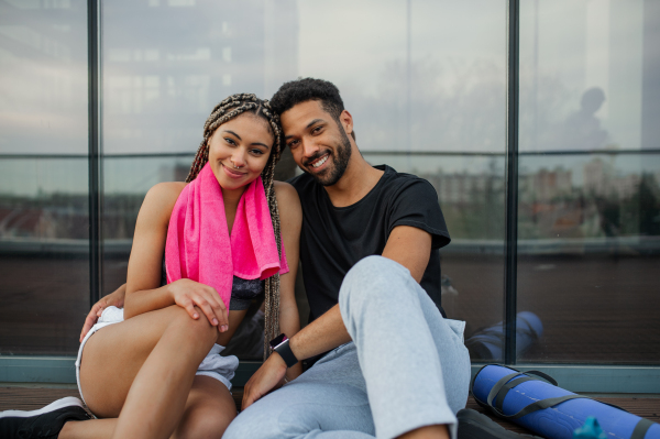 A young couple in love resting after exercise outdoors on terrace, looking at camera, sport and healthy lifestyle concept.